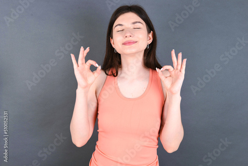 Caucasian woman wearing orange T-shirt over grey wall relax and smiling with eyes closed doing meditation gesture with fingers. Yoga concept.
