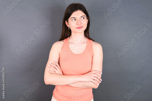 Pleased young beautiful Caucasian woman wearing orange T-shirt over grey wall keeps hands crossed over chest looks happily aside