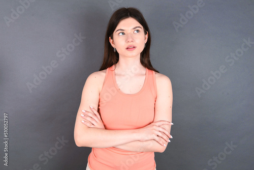 Charming thoughtful young beautiful Caucasian woman wearing orange T-shirt over grey wall stands with arms folded concentrated somewhere with pensive expression thinks what to do