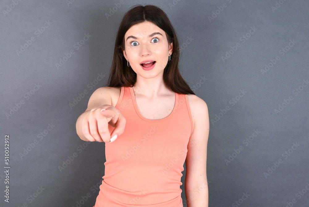 Excited positive young beautiful Caucasian woman wearing orange T-shirt over grey wall points index finger directly at you, sees something very funny. Wow, amazing