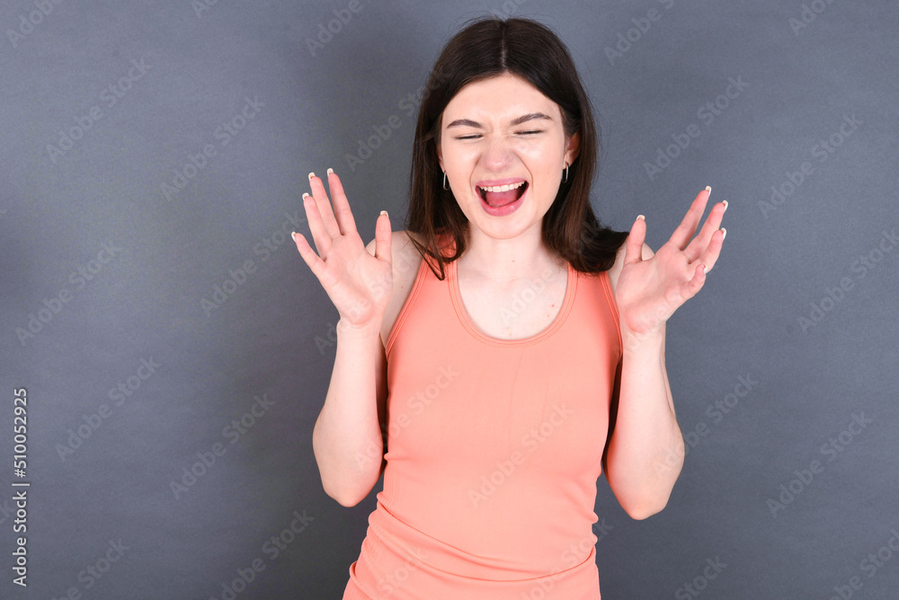 Crazy outraged Caucasian woman wearing orange T-shirt over grey wall screams loudly and gestures angrily yells furiously. Negative human emotions feelings concept Crazy outraged Caucasian woman wearing orange T-shirt over grey wall screams loudly and gestures angrily yells furiously. Negative human emotions feelings concept