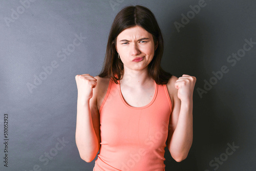 Irritated Caucasian woman wearing orange T-shirt over grey wall blows cheeks with anger and raises clenched fists expresses rage and aggressive emotions. Furious model © Roquillo