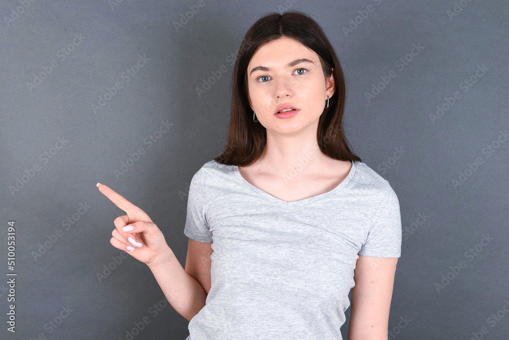 Serious young beautiful Caucasian woman wearing white T-shirt over grey wall smirks face points away on copy space shows something unpleasant. Look at this advertisement. Big price concept Serious young beautiful Caucasian woman wearing white T-shirt over grey wall smirks face points away on copy space shows something unpleasant. Look at this advertisement. Big price concept