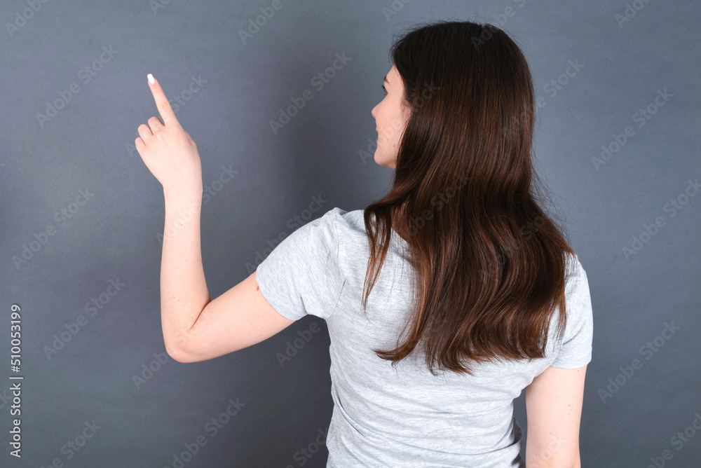 young beautiful Caucasian woman wearing white T-shirt over grey wall pointing to object on copy space, rear view. Turn your back young beautiful Caucasian woman wearing white T-shirt over grey wall pointing to object on copy space, rear view. Turn your back