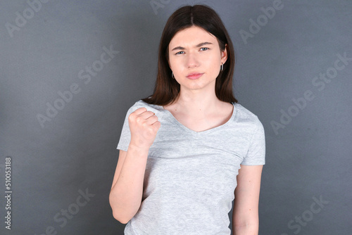  young beautiful Caucasian woman wearing white T-shirt over grey wall shows fist has annoyed face expression going to revenge or threaten someone makes serious look. I will show you who is boss