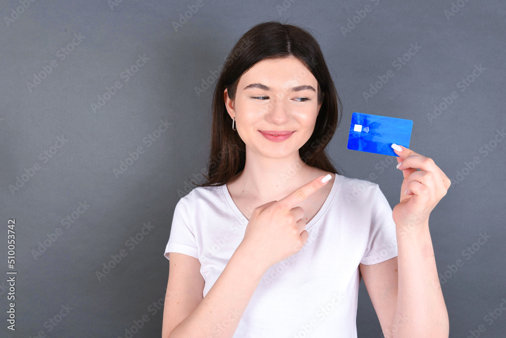 Beautiful caucasian girl standing over gray background and wearing white T-shirt, holding a credit card and pointing at it. Finance and online shopping concept. Beautiful caucasian girl standing over gray background and wearing white T-shirt, holding a credit card and pointing at it. Finance and online shopping concept.