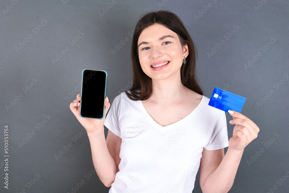 Young beautiful caucasian girl standing over gray background holding a credit card and an empty screen of smartphone. Advertisement and promotion concept. Young beautiful caucasian girl standing over gray background holding a credit card and an empty screen of smartphone. Advertisement and promotion concept.