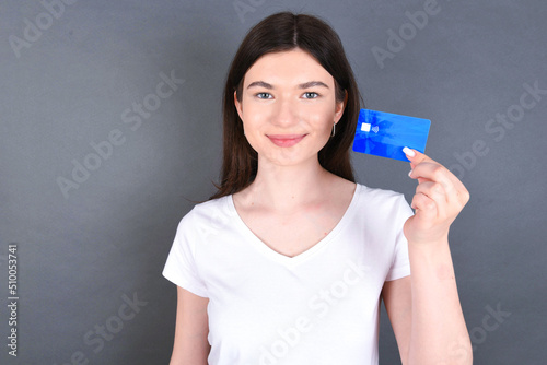Beautiful caucasian girl standing over gray background and wearing white T-shirt  holding a credit card and smiling to the camera. Finance and online shopping concept.