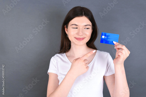 Beautiful caucasian girl standing over gray background and wearing white T-shirt  holding a credit card and pointing at it. Finance and online shopping concept.