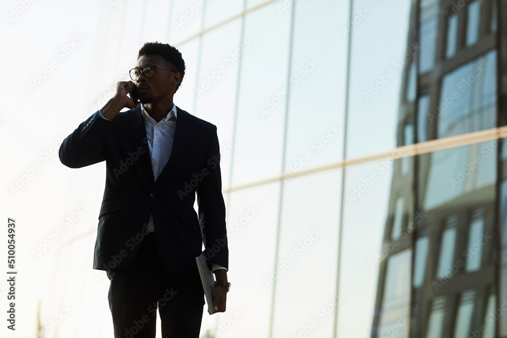 Pensive young black businessman in eyeglasses moving along street and communicating on mobile phone while leaving financial district Pensive young black businessman in eyeglasses moving along street and communicating on mobile phone while leaving financial district