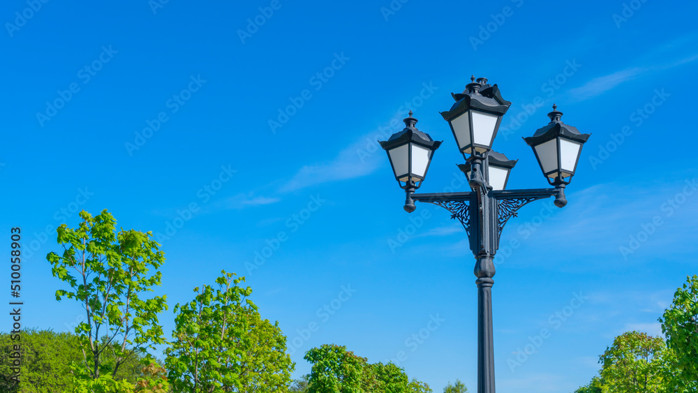 Vintage street lamp on blue sky and green trees background. Close-up. Old-fashioned lampost. Space for text.
