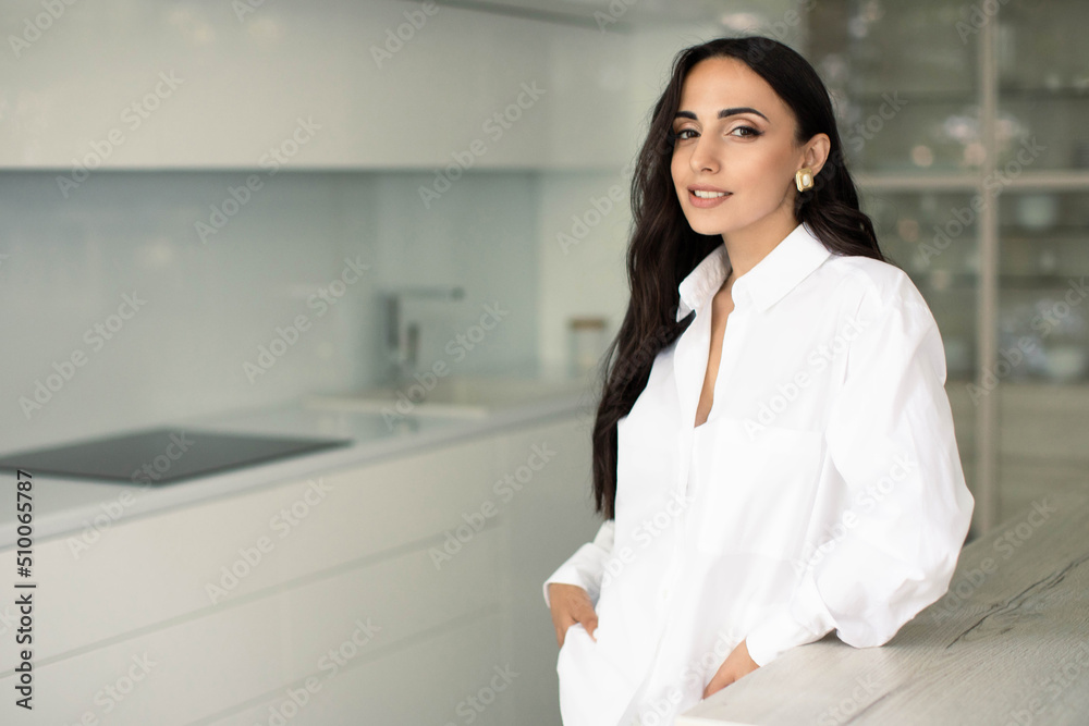 Brunette woman in white clothes standing indoors in apartment kitchen with design interior, smiling and confident. Freelancer or business woman at home office Brunette woman in white clothes standing indoors in apartment kitchen with design interior, smiling and confident. Freelancer or business woman at home office