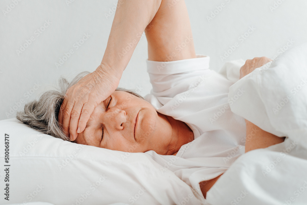 Lonely tired elderly woman with closed eyes holding her head while lying in bed in bedroom. Headache, insomnia, depression, women's health concept