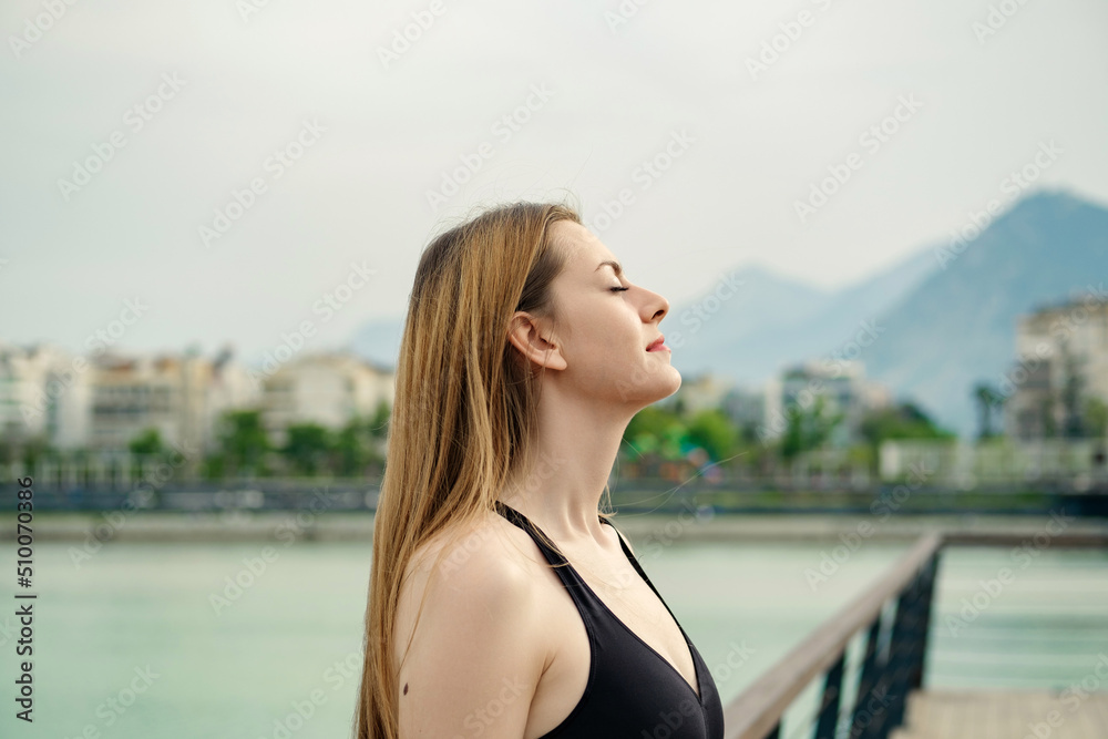 Young caucasian woman wearing black sports bra standing on city park, outdoors relaxing with eyes closed, feeling alive, breathing, dreaming.