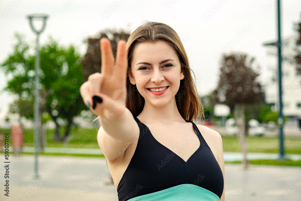 Young brunette girl smiling happy wearing sportive clothes on city park, outdoors showing and pointing up with fingers number two while smiling confident and happy. Selective focus on her face.