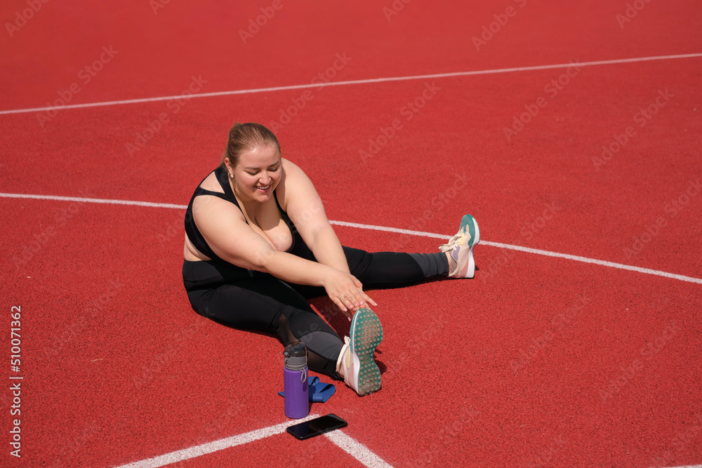Plump fat woman is doing sports on rubber covering of stadium. The person does stretching before fitness. Outdoor training on summer morning. Striving for weight loss , maintaining healthy lifestyle