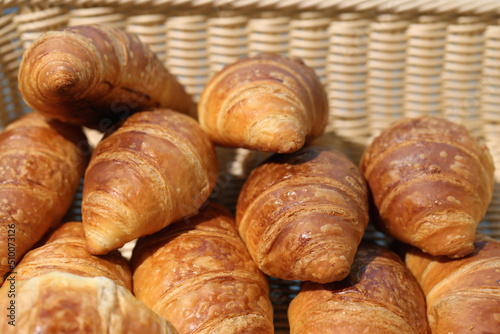 Big pile of multiple big and crunchy breakfast croissants in a wooden basket. Photographed outdoors during a sunny summer day. Delicious Parisian style breakfast bread. Closeup color image.