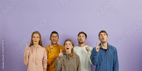 Group of different mixed race multiethnic people struck by amazing idea and bright solution standing on color background and pointing index fingers up with open mouths and surprised facial expressions