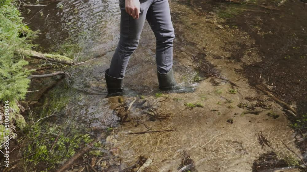 Unrecognizable slim young man in rubber boots walks along riverbed of cool clean stream, close-up, slow motion. Guy on hike crosses river ford along sandy bottom. Walk, rest and relax in wild nature. Unrecognizable slim young man in rubber boots walks along riverbed of cool clean stream, close-up, slow motion. Guy on hike crosses river ford along sandy bottom. Walk, rest and relax in wild nature.