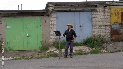 A man in a straw hat holds a shovel against the backdrop of garages. Dressed in a black shirt and pants. Bearded. warm day. worker. builder. Illegal migrant. Searching for a job © Evgeniy Trofimenko