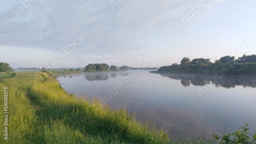 Morning on the river. Light fog and soft sunshine. Tall grass and trees grow along the banks and are reflected in the calm water. There is a slight cloudiness in the sky