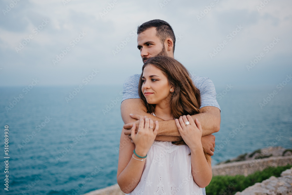 A Hispanic man is hugging from behind his brunette girlfriend in the park in Spain in the evening. A couple of tourists are staring at the sea on a date at the sunset in Valencia. A Hispanic man is hugging from behind his brunette girlfriend in the park in Spain in the evening. A couple of tourists are staring at the sea on a date at the sunset in Valencia.