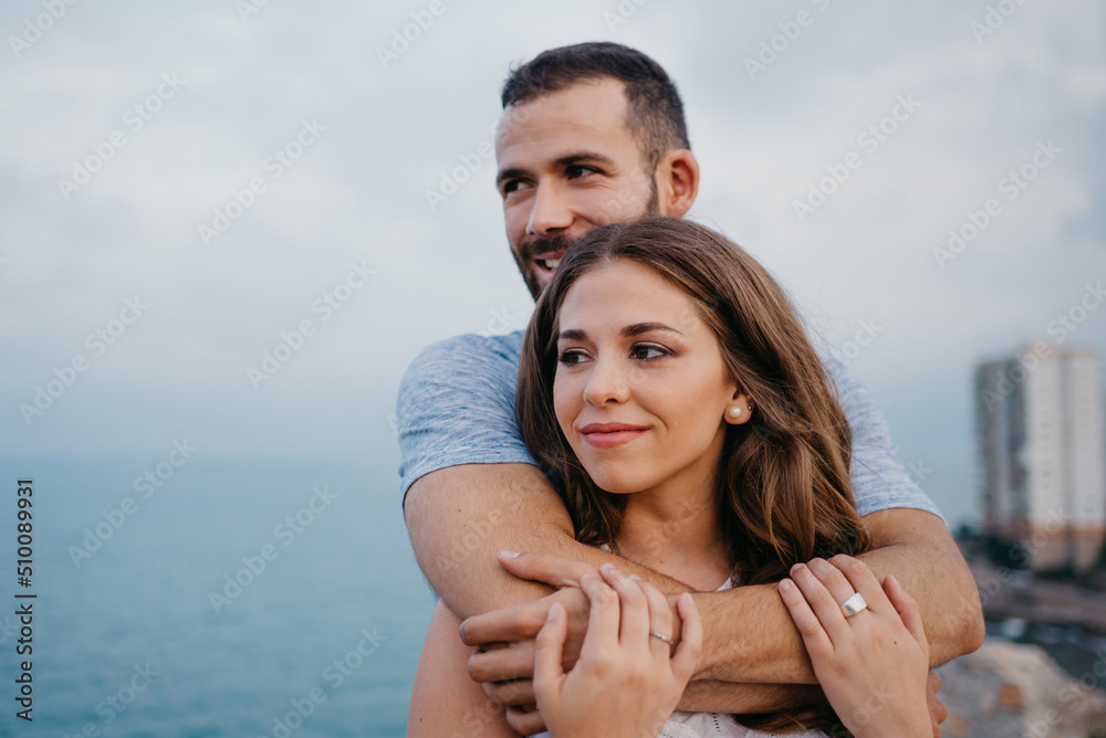 A close photo of a Hispanic man who is hugging from behind his brunette girlfriend in the park in Spain in the evening. A couple of tourists are staring at the sea on a date at the sunset in Valencia. A close photo of a Hispanic man who is hugging from behind his brunette girlfriend in the park in Spain in the evening. A couple of tourists are staring at the sea on a date at the sunset in Valencia.