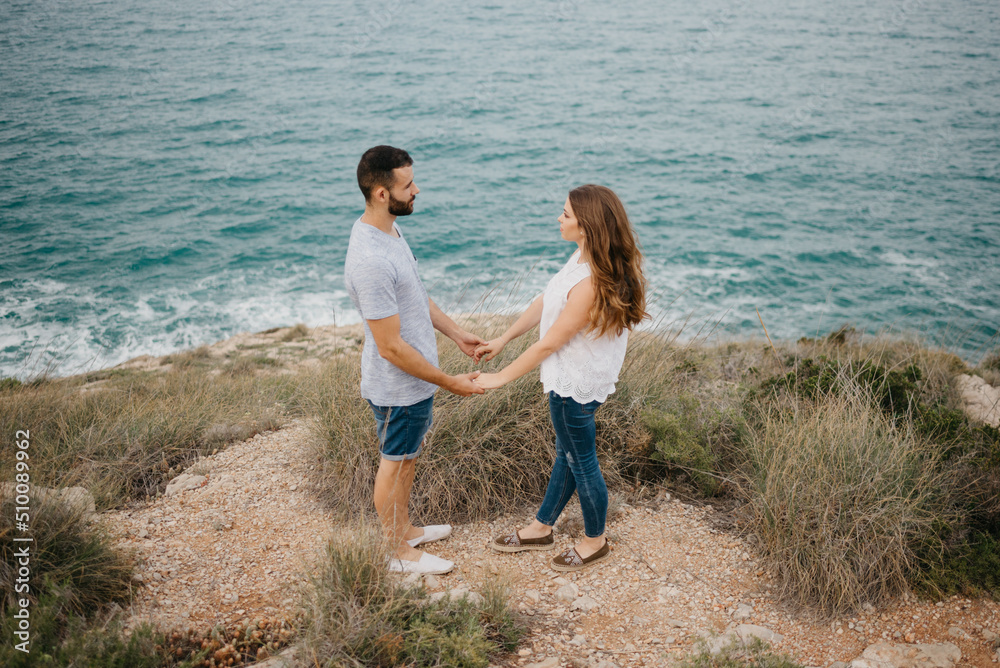 A Hispanic man is holding hands with his Latina girlfriend with the sea in the background in the highland park in Spain. A couple of tourists are enjoying each other near the sea at sunset in Valencia A Hispanic man is holding hands with his Latina girlfriend with the sea in the background in the highland park in Spain. A couple of tourists are enjoying each other near the sea at sunset in Valencia