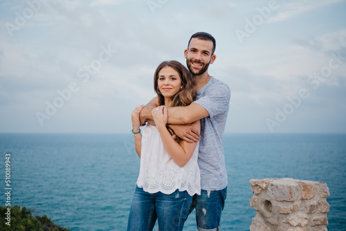 A Hispanic man is hugging from behind his brunette girlfriend in the park in Spain in the evening. A couple of tourists are posing near the sea on a date at the sunset in Valencia.