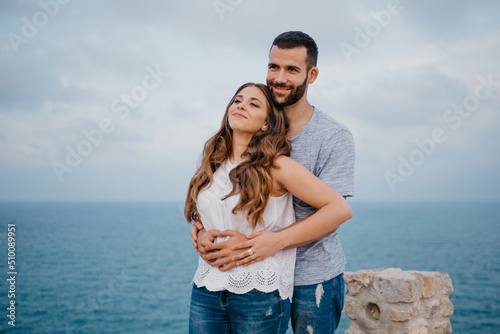 A Hispanic man is hugging from behind his happy girlfriend in the highland park in Spain in the evening. A couple of tourists are enjoying each other near the sea on a date at sunset in Valencia.