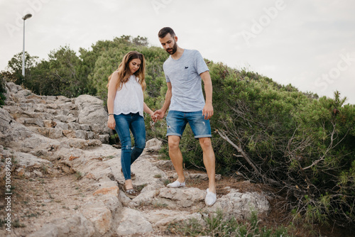 A Hispanic man is helping his tired wife descend the ancient stone stairs in the highland park in Spain. A couple of tourists are enjoying hiking with each other on a date at sunset in Valencia.