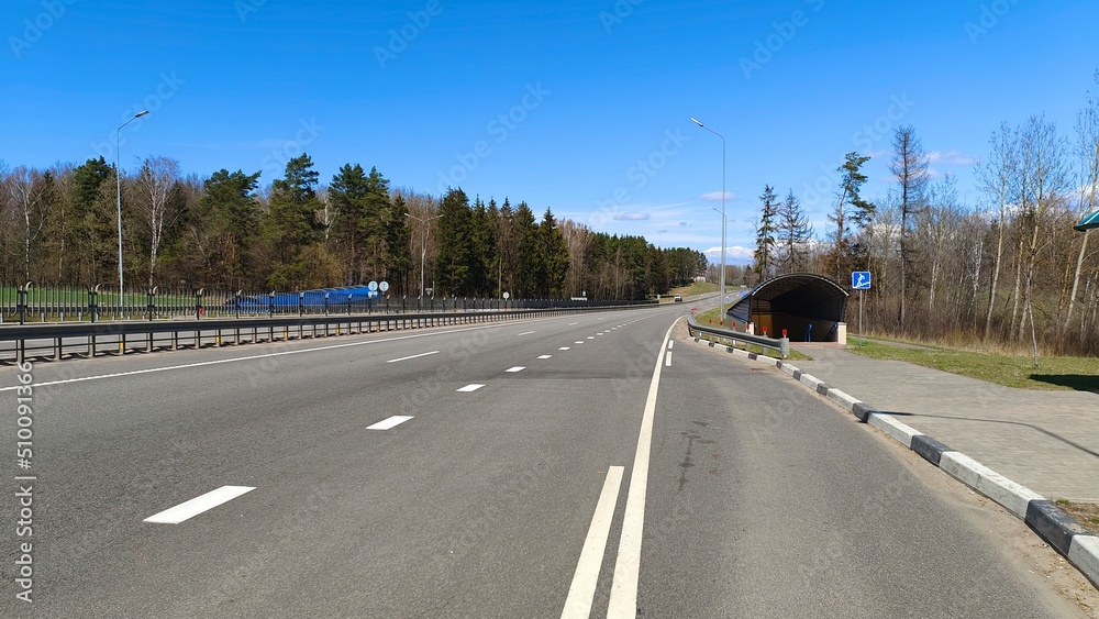 The paved motorway with markings is equipped with side and central metal fences, poles with lighting lights and an underground pedestrian crossing. There is a forest growing along the road