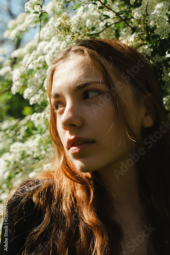 vertical photo of a beautiful red-haired woman sitting near a bush of white flowers looking pleasantly to the side  illuminated by the rays of the setting sun