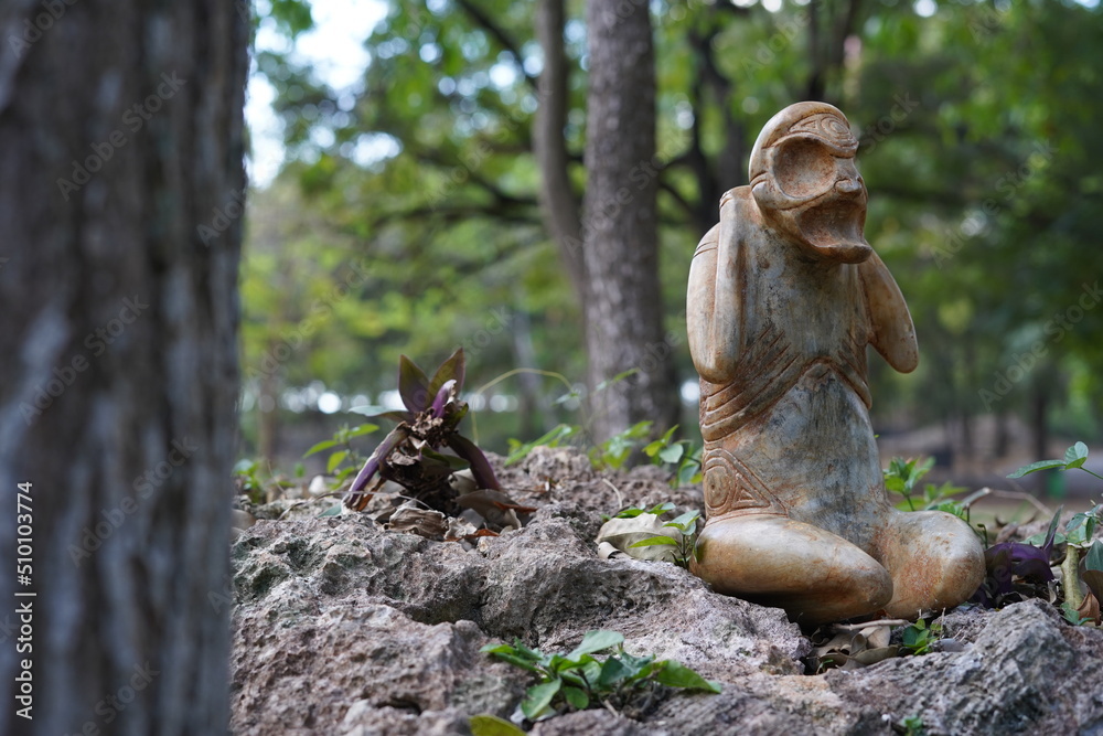Taino Antique Stone Cemi Idol God Figure standing on rocks on the ground next to a tree, close up. Taino Mythology. Taino Antique Stone Cemi Idol God Figure standing on rocks on the ground next to a tree, close up. Taino Mythology.