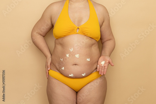 Fat woman in yellow swimsuit apply spot white body cream on thick sagging stomach, beige background. Slimming, fighting overweight, obesity and cellulite. Plus size people and body positive concept.
