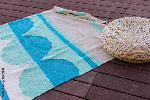 Blue and teal blanket sits on deck outside, next to woven poof
 photo