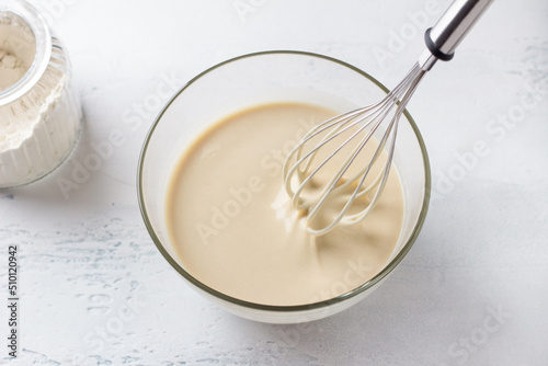A glass bowl of dough for Finnish pancakes with a whisk and a jar of flour on a light gray background. Cooking delicious homemade food