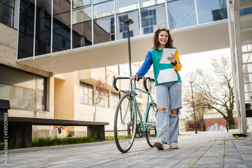 One woman young adult caucasian female walking by the university building holding bicycle and digital tablet in day real people copy space happy joyful full length front view