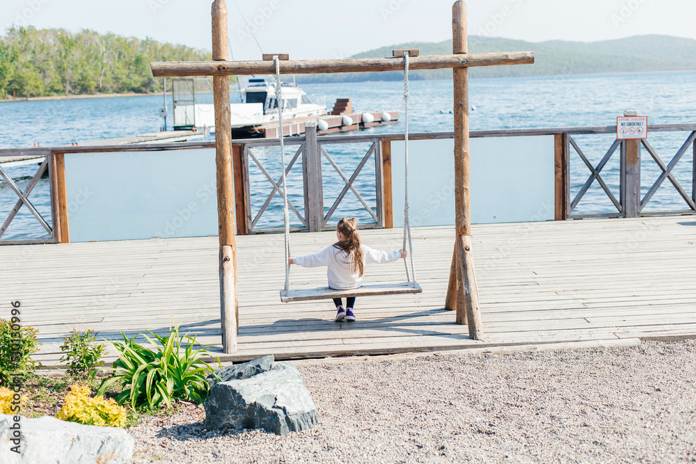 A little girl swings on a wooden swing by the sea. Back view. Outdoor activities A little girl swings on a wooden swing by the sea. Back view. Outdoor activities