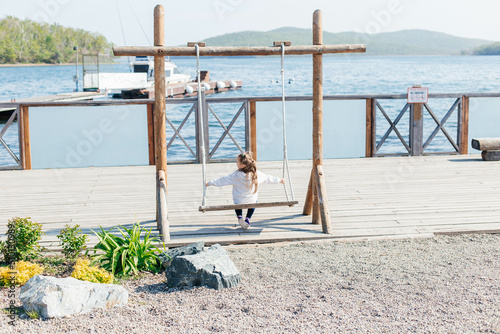 A little girl swings on a wooden swing by the sea. Back view. Outdoor activities