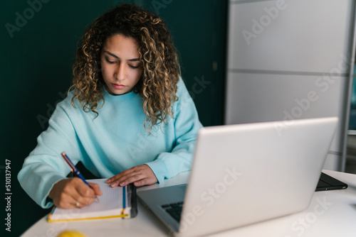 Focused woman writing in notepad sitting at laptop