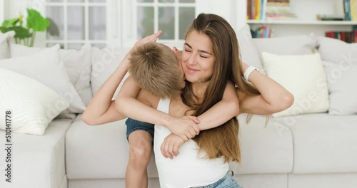 Young mother and little son have fun at home in the living room. Mom hugs and kisses her son. Active family, parent and child spend time together enjoying fun activities.
