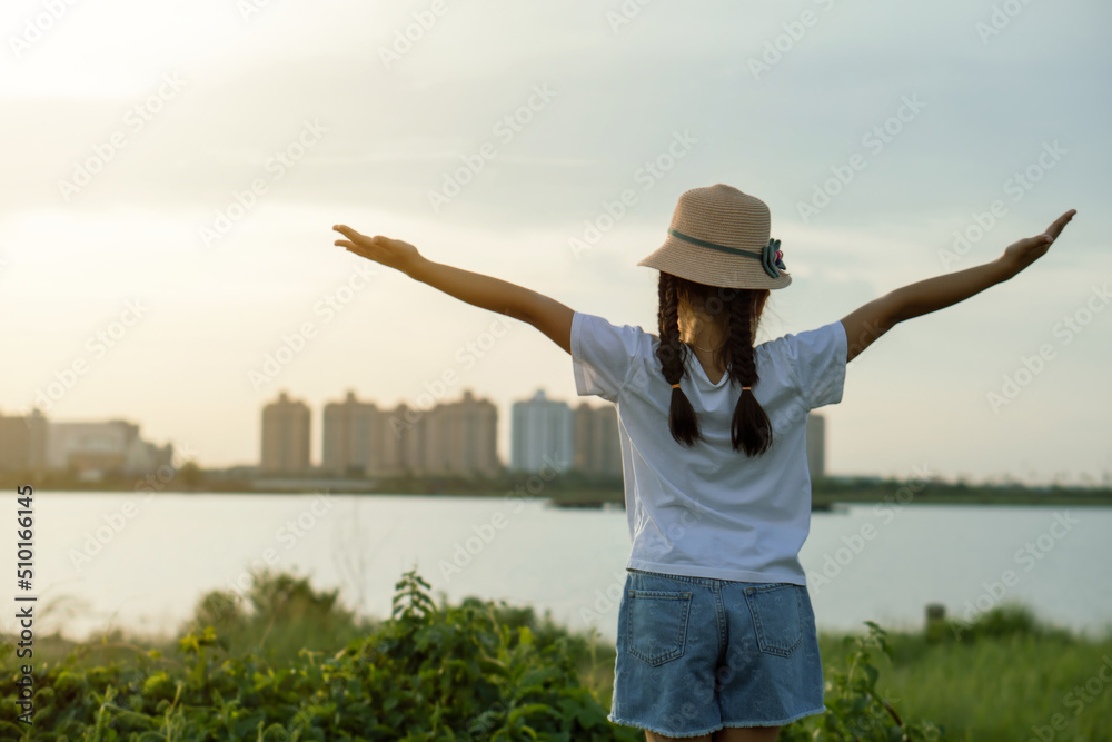 Asian kid girl wearing hat raising arms relaxing feeling happy looking at sunset on sky enjoying sunlight and Thatluang lake view in Vientiane city, Laos during vacation, freedom hope concept.