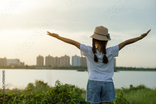 Asian kid girl wearing hat raising arms relaxing feeling happy looking at sunset on sky enjoying sunlight and Thatluang lake view in Vientiane city, Laos during vacation, freedom hope concept. photo