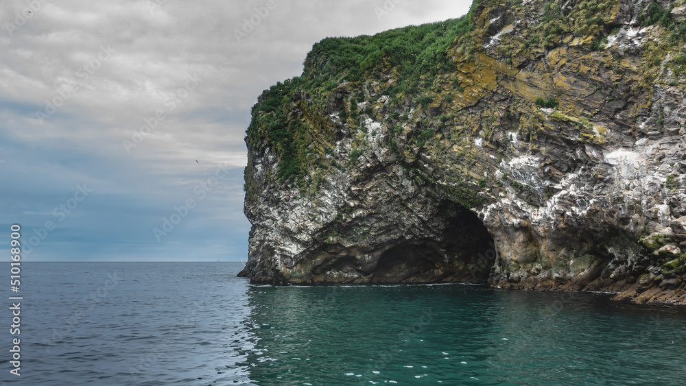 An island in the Pacific Ocean. Seabirds nest on steep rocky slopes with sparse vegetation. The entrance to the dark grotto is visible above the surface of the water. Clouds in the sky. Kamchatka.