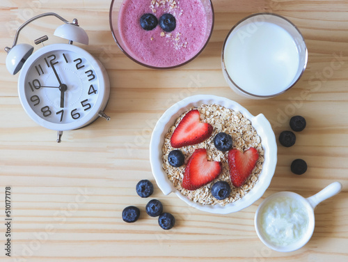 flat lay of alarm clock, breakfast with oat or granola in white bowl, fresh blueberries, strawberries, a glass of milk, blueberry smoothie and yogurt on wooden table. Healthy breakfast concept.