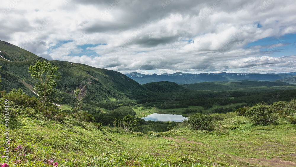 View of the lake from the mountain slope. The meadow has lush green vegetation. The path goes down. Picturesque clouds in the blue sky. Reflection. Kamchatka. Vachkazhets. Lake Tahkoloch