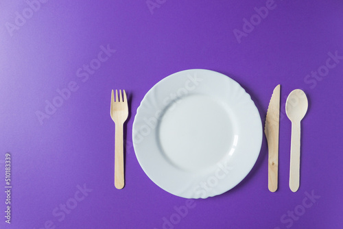 Tableware from natural materials - bamboo wooden fork, spoon and knife with white plate isolated on a violet background.