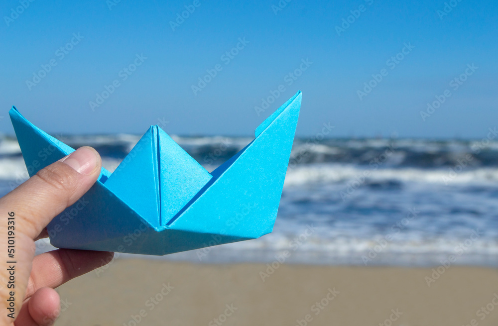 Person holding blue paper boat in hand and playing floating in air on background sea waves. Blue sky and sand beach on sunny summer day Concept tourism vacation holiday rest dreaming traveling sailing
