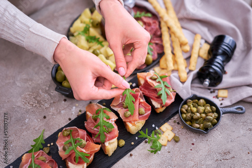 woman puts arugula leaves to prosciutto ham bruschetta with traditional antipasto meat plate on background